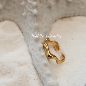 May include: Gold-toned ring with a wavy, abstract design. The ring is on a white, sandy surface. Hint Fine Jewelry is written in the background.
