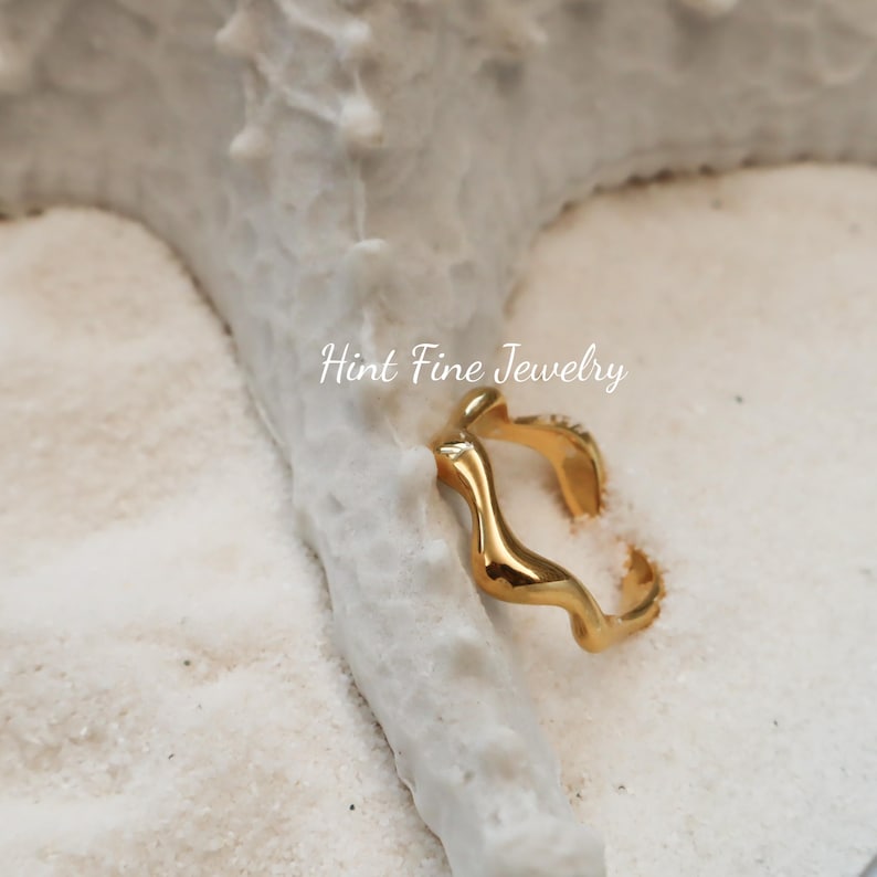 May include: Gold-toned ring with a wavy, abstract design. The ring is on a white, sandy surface. Hint Fine Jewelry is written in the background.