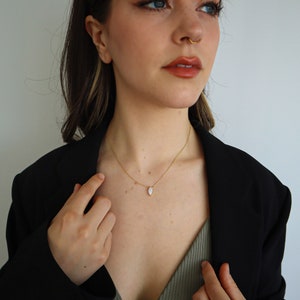 May include: A gold necklace with a small, teardrop-shaped pendant. The pendant is white and has a gold border. The necklace is worn over a green top and a black blazer.