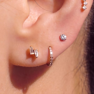 May include: A gold ear piercing set featuring a small stud with two baguette-cut clear stones, a thin gold hoop earring encrusted with small clear stones, a single round clear stone stud, and a three-stone curved gold stud at the top.