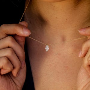 May include: A delicate gold necklace featuring a marquise-cut diamond pendant. The diamond is set in a simple, elegant design, suspended from a fine gold chain. The necklace is shown against a dark background, highlighting the sparkle of the diamond.
