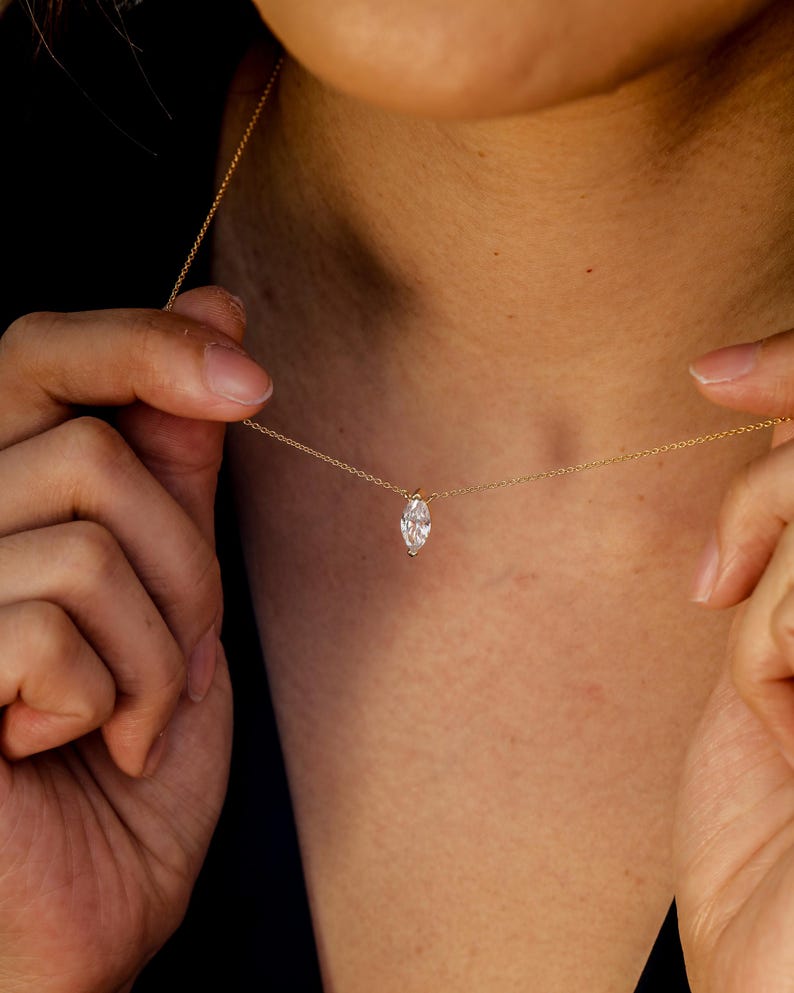 May include: A delicate gold necklace featuring a marquise-cut diamond pendant. The diamond is set in a simple, elegant design, suspended from a fine gold chain. The necklace is shown against a dark background, highlighting the sparkle of the diamond.