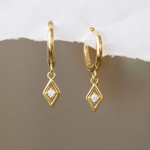 May include: A pair of gold-coloured hoop earrings. Each earring has a diamond-shaped charm with a small, round, clear gemstone in the centre. The earrings are displayed against a neutral background.