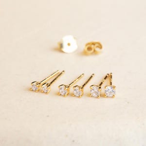 May include: Gold-colored stud earrings with clear, round gemstones. The earrings are displayed with their backs, showcasing a delicate and elegant design. The image also includes a pair of earring backs.