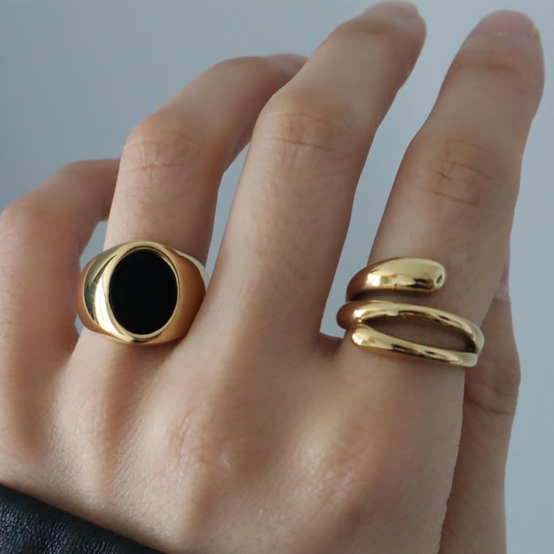 May include: Two gold rings on a hand. One ring has a black oval stone, the other is a twisted band.
