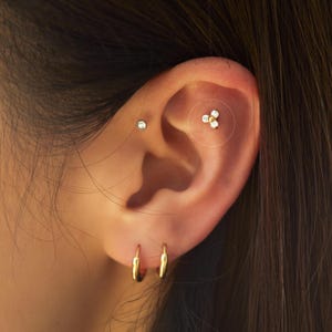 May include: Close-up of an ear with multiple piercings. The ear has a small, round stud, a small gold hoop earring, and a cluster of three small, clear stones with a gold centre. The jewellery is gold-toned.