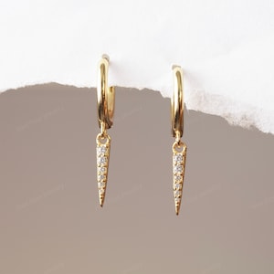 May include: A pair of gold-coloured hoop earrings with a dangling, pointed charm. Each charm is adorned with small, clear gemstones. The earrings are displayed against a neutral background, highlighting the jewellery's design.