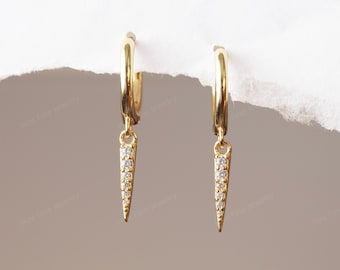 May include: A pair of gold-colored hoop earrings with a dangling, pointed charm. Each charm is adorned with small, clear gemstones. The earrings are displayed against a neutral background, highlighting the jewelry's design.