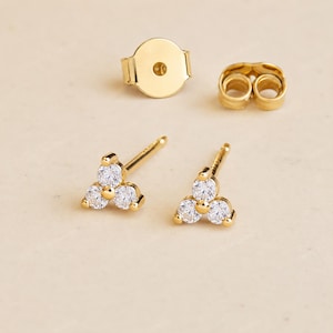 May include: A pair of gold-colored stud earrings, each featuring three clear, round gemstones arranged in a triangular cluster. The image also shows a gold earring back and a butterfly earring back.