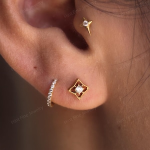 May include: A gold ear piercing set featuring a four-pointed star stud with a central clear stone, a square-shaped stud with an openwork frame and a central clear stone, and a small gold hoop earring lined with tiny clear stones.