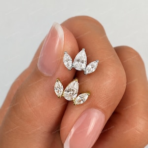 May include: Two crown-shaped stud earrings, one in silver-toned metal and one in gold-toned metal, each featuring three clear, pear-shaped faceted stones arranged in a fan-like cluster, showcasing a brilliant sparkle and polished metal settings.
