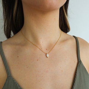 May include: A gold-colored necklace with a marquise-cut gemstone pendant. The pendant is a clear, sparkling stone set in a gold-colored bezel. The necklace is worn on a person's neck, against a green tank top.