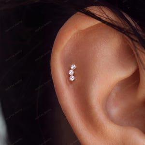 May include: A silver-toned cartilage earring featuring three small, clear, round-cut stones arranged in a vertical, slightly curved line. The jewellery is positioned on the upper ear cartilage, showcasing a delicate, minimalist design with a polished metal finish.