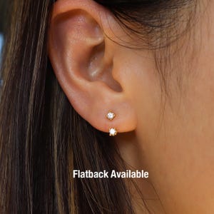 May include: Close-up of two small, gold-colored stud earrings, each featuring a clear, sparkling gemstone. The earrings are set in a four-prong setting. The text "Flatback Available" is visible at the bottom of the image.