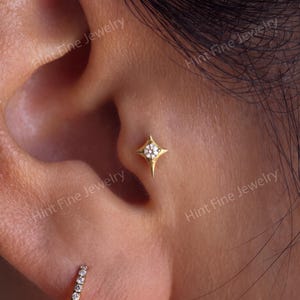 May include: A gold tragus earring featuring a four-pointed star shape with a central round clear gemstone, accompanied by a small gold hoop earring with a row of tiny clear stones set along the lower edge of the earlobe.