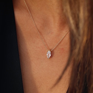 May include: A delicate gold necklace featuring a marquise-cut diamond pendant. The diamond is set in a gold bezel and hangs from a fine gold chain. The necklace is shown against a dark background.