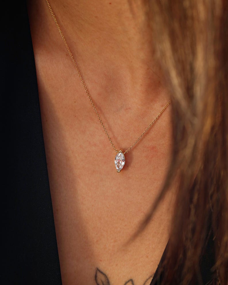 May include: A delicate gold necklace featuring a marquise-cut diamond pendant. The diamond is set in a gold bezel and hangs from a fine gold chain. The necklace is shown against a dark background.