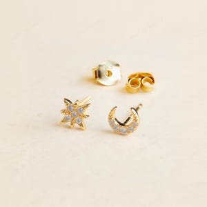 May include: A pair of gold-colored stud earrings, one in a starburst design and the other in a crescent moon shape. Both earrings are embellished with small, clear gemstones. Also included are two earring backs.