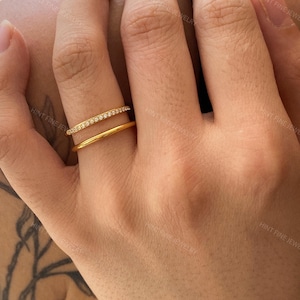 May include: Two gold rings are displayed on a hand. One ring is a simple band, while the other features a row of small, clear stones. The rings are set against the skin, with the words "HINT FINE JEWELRY" visible in the background.