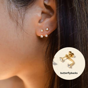 May include: A gold-toned earring featuring two clear, round-cut stones set in a prong setting. The design includes a curved post that connects the two stones, allowing them to appear as two separate piercings. The earring includes butterfly backs.