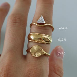 Peut inclure: Trois bagues dorées sont présentées sur un doigt. Le style A présente une pierre transparente triangulaire. Le style B est une large bande avec un motif de corde. Le style C a un motif en forme d'éventail. Le texte "Style A", "Style B" et "Style C" est visible.
