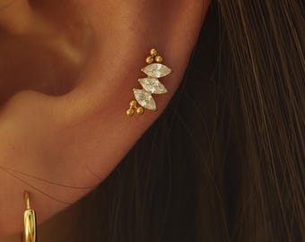 May include: Gold-coloured ear jewellery featuring a cluster of small, round beads and marquise-cut, clear gemstones. A small gold hoop earring is also visible. The jewellery is set against the backdrop of a person's ear and hair.