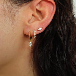 May include: A close-up of a person's ear with three gold earrings. The earrings are all set with small, clear stones. The earrings are a hoop, a bar with three stones, and a single stone.
