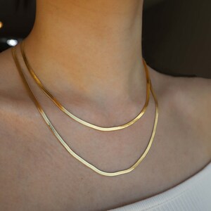 May include: Two gold chain necklaces layered on a person's neck. The chains are thin and have a snake chain design.