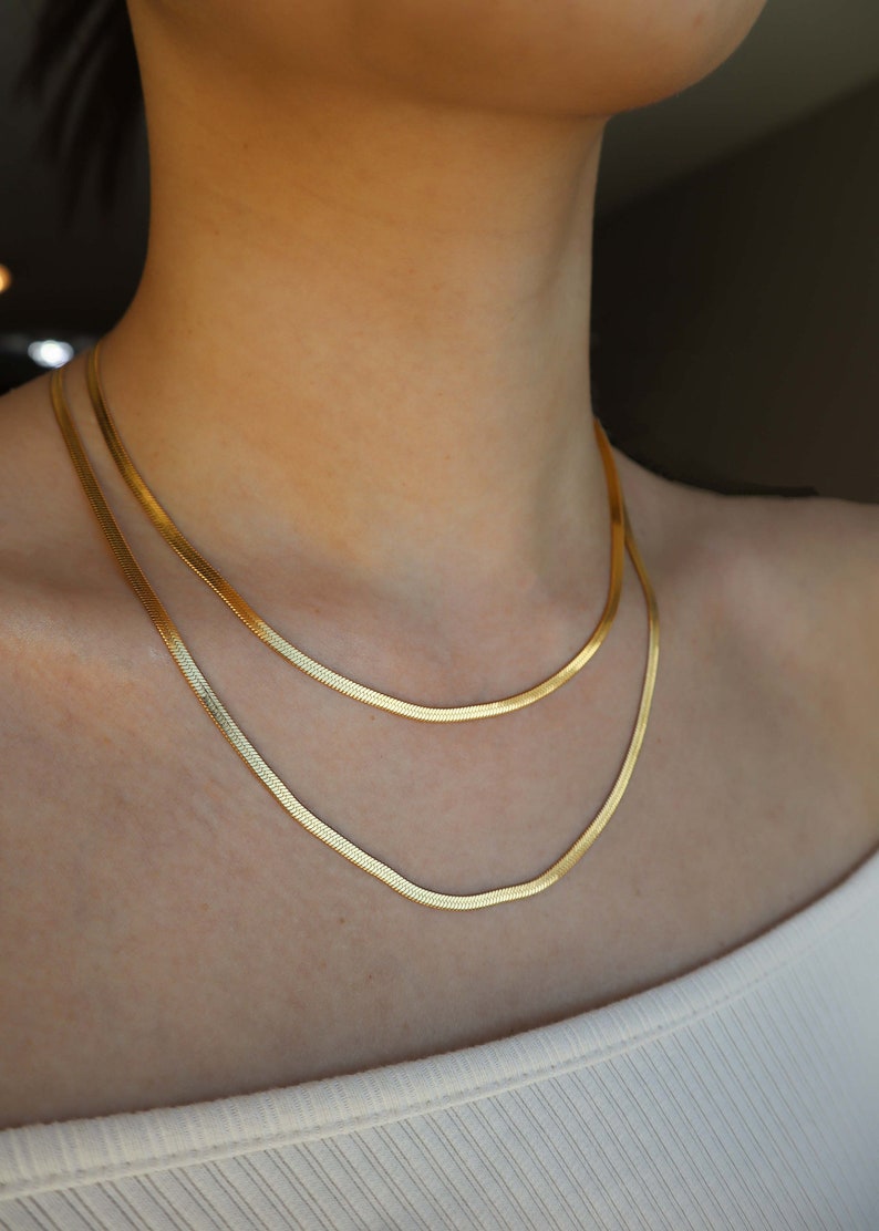 May include: Two gold chain necklaces layered on a person's neck. The chains are thin and have a snake chain design.