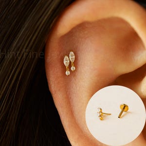 Peut inclure: Bijou de piercing d'oreille doré avec deux cristaux transparents en forme de marquise et un cristal rond pendant. Le bijou est présenté sur une oreille, avec un poteau et un fermoir d'oreille séparés dans un encart circulaire.