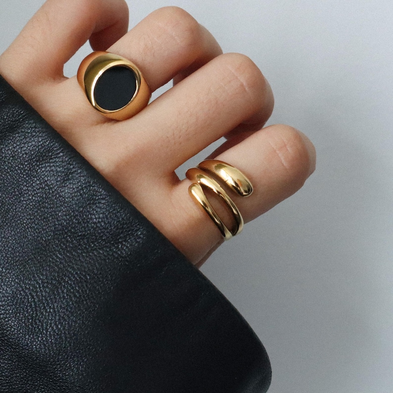 May include: Two gold rings on a hand. One ring is a simple band, the other is a thick oval ring with a black stone.