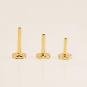 May include: Three gold-colored, cylindrical jewelry posts with flat, circular bases. The posts are of varying heights, arranged in a row against a neutral background. The posts have a polished, reflective surface, suggesting a metallic material.