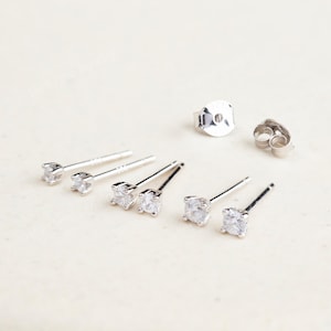 May include: A collection of silver stud earrings with clear, round cubic zirconia stones. The earrings are displayed with their backs, showcasing the post and butterfly clutch closures. The jewelry is set against a neutral background.