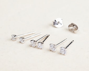 May include: A collection of silver stud earrings with clear, round cubic zirconia stones. The earrings are displayed with their backs, showcasing the post and butterfly clutch closures. The jewelry is set against a neutral background.