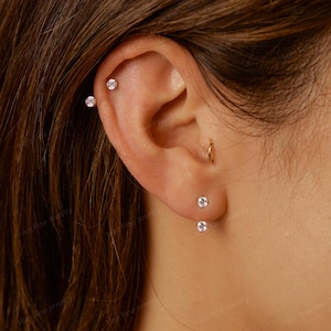 May include: Close-up of an ear with multiple pieces of jewellery. There are three small, round, clear stud earrings and a small gold hoop earring. The earrings are set in a light-coloured metal. The ear is against a blurred background.