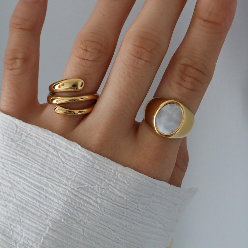 May include: Two gold rings on a hand. One ring is a simple oval with a white stone. The other ring is a twisted band.