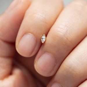 May include: A small, gold-colored earring with a marquise-cut, clear gemstone. The earring is held between fingers, showcasing its delicate design and sparkling stone. The earring is a stud style, perfect for everyday wear.