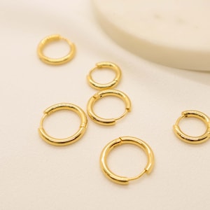May include: Five gold hoop earrings of different sizes on a white surface.