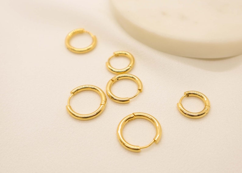 May include: Five gold hoop earrings of different sizes on a white surface.