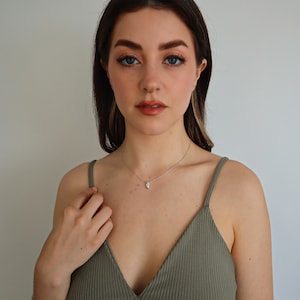 May include: A silver necklace with a teardrop-shaped pendant. The pendant is a clear, sparkling stone. The woman in the image is wearing a green spaghetti strap top.