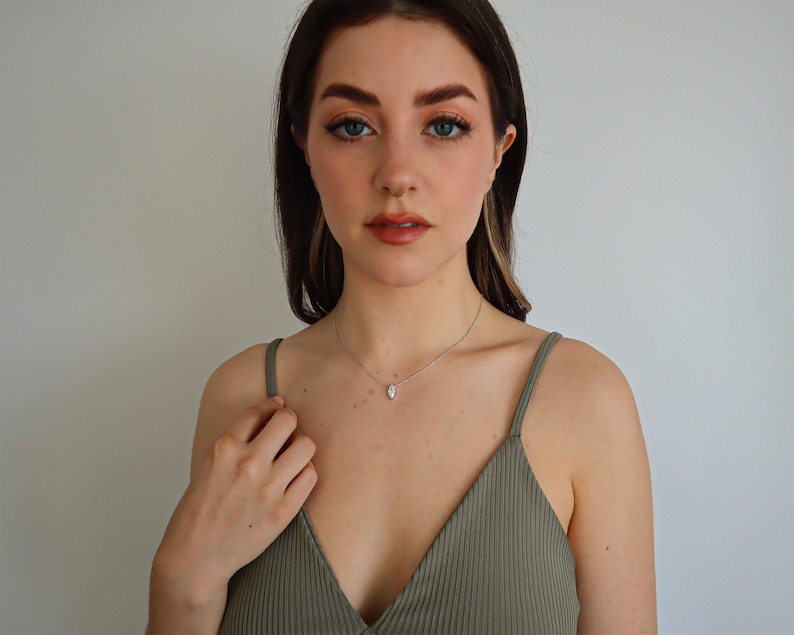 May include: A silver necklace with a teardrop-shaped pendant. The pendant is a clear, sparkling stone. The woman in the image is wearing a green spaghetti strap top.