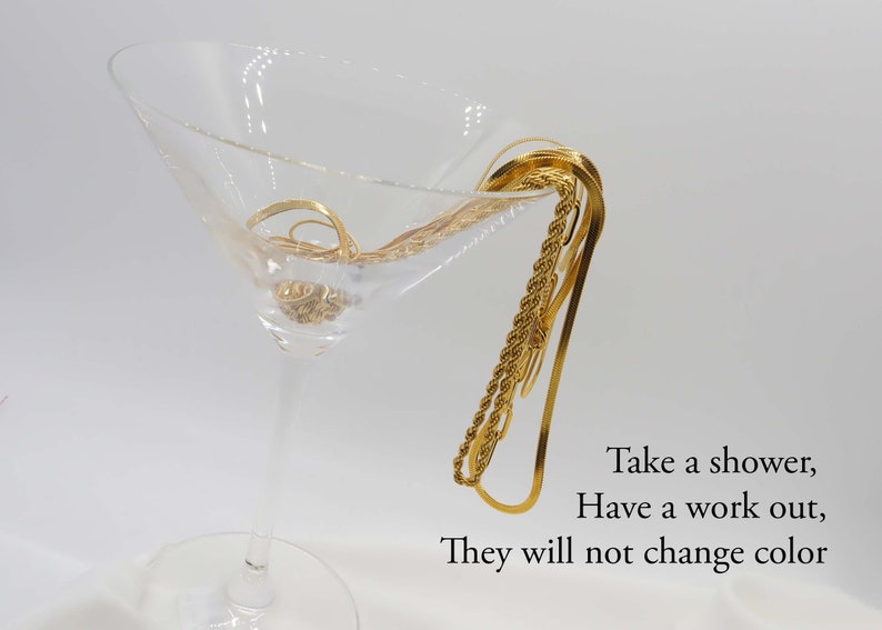 May include: A close-up of three gold chains in a martini glass. The chains are made of different styles of gold links. The text "Take a shower, Have a work out, They will not change color" is visible below the glass.