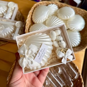 Può includere: Bomboniere a forma di conchiglia bianca in scatole trasparenti, legate con nastri bianchi. Le scatole contengono un oggetto a forma di conchiglia, piccoli oggetti rotondi e un piccolo oggetto quadrato. Il testo sull'etichetta recita "with love ZEYNEP & PAWEL 10.04.2023".