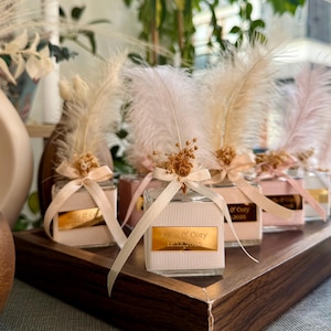 Può includere: Una collezione di piccoli vasetti quadrati di vetro con dettagli rosa e bianchi, ognuno adornato con una piuma e un nastro. I vasetti sono disposti su un vassoio di legno scuro, con etichette dorate che riportano "Kelly & Cory" e una data.