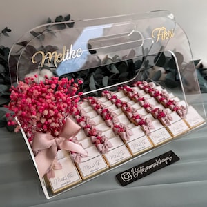 May include: A clear acrylic box with a handle, filled with small white boxes wrapped in pink ribbon and pink flowers. The box is decorated with the names "Melike" and "Fikri" in gold lettering. The Instagram handle "@butikcarminhikayesi" is printed on a black tag.