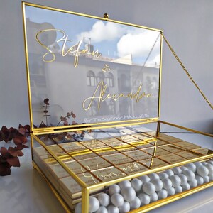 May include: A gold glass box with a hinged lid and a chain. The lid has the names "Stefan & Alexander" written in gold script. The box is filled with small gold-wrapped chocolates and white pearl-like sweets.