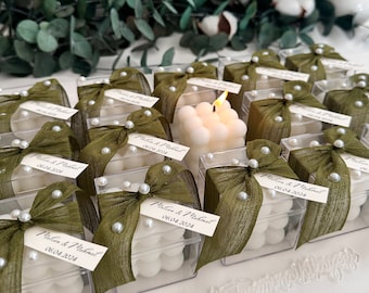 May include: A collection of small, clear boxes with white candles inside. Each box is decorated with a green ribbon and white pearl beads. The boxes have a label with the text "Melisa & Michael 06.04.2024".