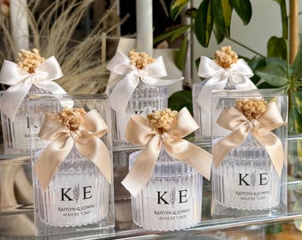 May include: Several small, clear glass jars with ribbed sides are arranged in a display. Each jar is topped with a satin bow and a small dried floral accent. The jars have a label with the initials "KE" and the names "Kaitlyn & Edwin" and the date "August 7, 2025".