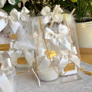 May include: A clear acrylic display stand holding a white candle in a ribbed glass jar, adorned with a white satin ribbon bow, white feathers, and a gold-toned metal tag labeled Amira. A thin gold chain hangs over the front of the display.