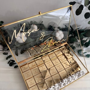 May include: A gold glass box with a gold chain handle filled with small gold boxes that say "Mosa & Zhraa" and white candy. The lid of the box has the names "Mosa & Zhraa" written in gold cursive.